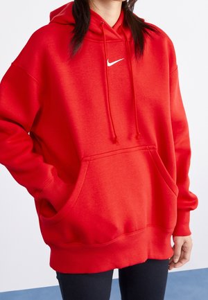 Persoon met een felrode Nike-hoodie met trekkoorden en een voorzak, staat met één hand in de zak.