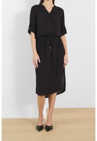 Czarna sukienka do kolan z dekoltem w serek, zwijanymi krótkimi rękawami, ściągniętą w talii i bocznymi rozcięciami. Wykonana z lekkiego materiału. Noszona z czarnymi szpilkami.