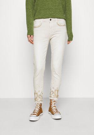 Personne portant un jean slim blanc avec broderie beige paisley et florale près des chevilles, associée à des baskets montantes marron et un pull vert.