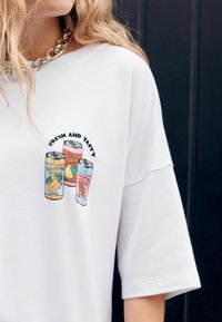 Fehér, túlméretezett póló, amelyen három színes italdoboz grafikai mintája látható, a "FRESH AND TASTY" felirattal fölötte.