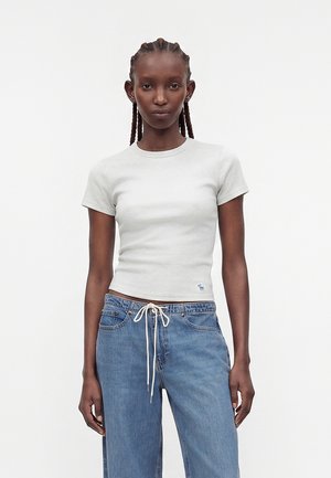 Donna con capelli intrecciati che indossa una maglietta bianca aderente a maniche corte e jeans blu larghi con coulisse bianca, in piedi davanti a uno sfondo semplice.