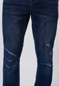 Jeans de mezclilla azul oscuro con un corte ajustado, detalles desgastados con roturas deshilachadas en ambas rodillas y costuras de contraste.