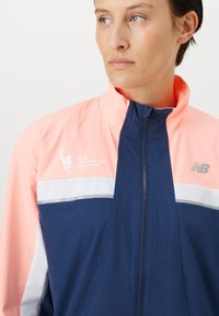Chaqueta de nailon azul marino y rosa con cierre de cremallera, con rayas blancas y plateadas y un logo del Maratón de Nueva York 2025.