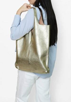 Metallic gold Tragetasche mit glatter Textur, großem Körper und betonten Nähten. Über der Schulter getragen, kombiniert mit einem blauen Oberteil und weißen Hosen.