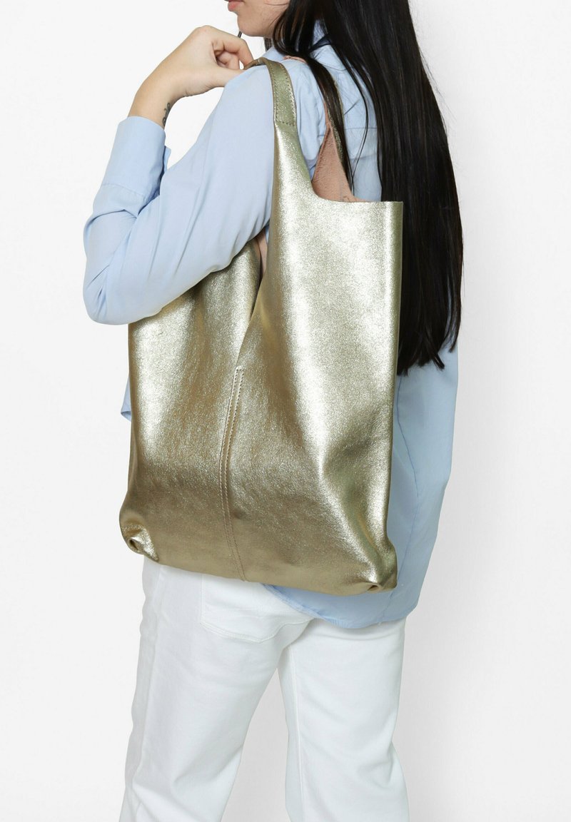 Borsa a tracolla metallica dorata con una texture liscia, grande corpo e cuciture accentuate. Indossata sulla spalla, abbinata a una camicia blu e pantaloni bianchi.