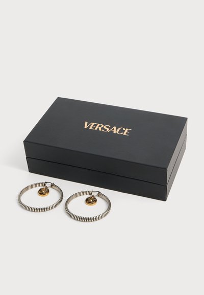 Paire de boucles d'oreilles créoles en argent avec breloques tête de lion en or à côté d'une boîte à bijoux Versace noire sur fond blanc.