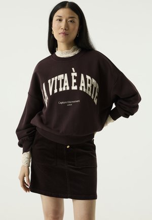 Vrouw met lang zwart haar draagt een donkere sweater met de tekst “La Vita È Arte”, kanten coltrui en bijpassend kort rokje tegen een effen achtergrond.