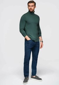 Grünes langärmliges Rollkragenoberteil, kombiniert mit dunkelblauen Jeans, ergänzt durch graue Schuhe und eine silberne Uhr, vor einem neutralen Hintergrund.