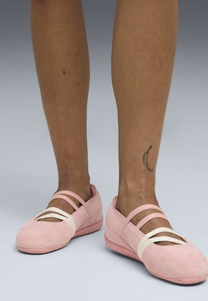 Pieds portant des chaussures de style ballet roses avec deux brides roses et une blanche sur chaque pied, tatouage de croissant de lune visible à la cheville de la jambe droite.