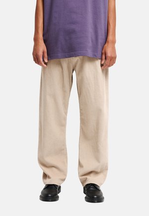 Person trägt beige weite Cordhosen, schwarze Schuhe und ein lila Hemd, gezeigt von der Taille bis zu den Füßen vor einem einfarbigen Hintergrund.