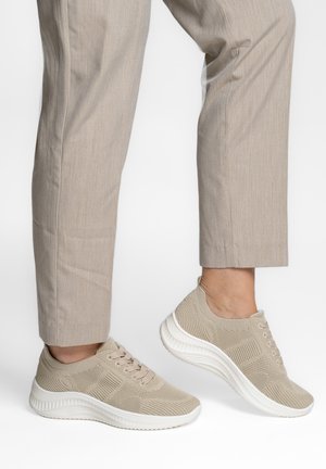 Persona con zapatillas de punto beige con suelas blancas y pantalones beige, con un pie levantado como si estuviera caminando o dando un paso adelante.