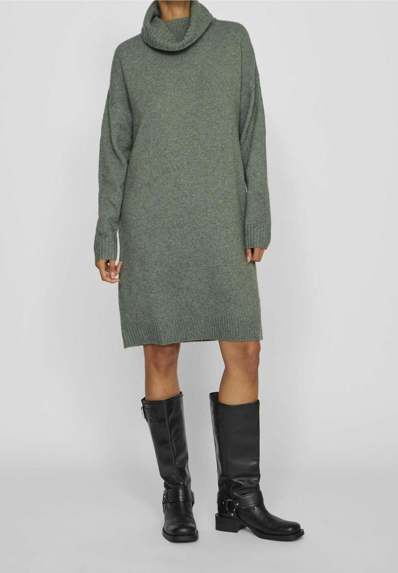 Personne portant une robe-pull longue grise à col roulé assortie à des bottes en cuir noires mi-mollet avec des boucles sur un fond gris.