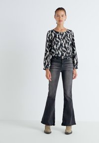Zwart-wit gestreepte blouse met pofmouwen, gecombineerd met nauwsluitende donkere jeans en beige enkellaarsjes, staande tegen een lichte achtergrond.