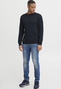 Mørkeblå strikkegenser med et teksturert mønster, båret med lyseblå jeans og marineblå joggesko. Har ribbestrikkede mansjetter og rund hals.