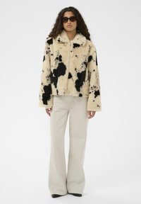 Jakna iz umetnega krzna v bež barvi s črnim kravjim vzorcem, z ovratnikom in zadrgo. Kombinirana z svetlobéž širokimi hlačami in črnimi čevlji.