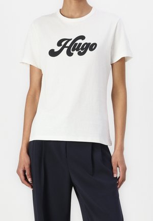 Hvid T-shirt med korte ærmer med sort "Hugo" tekst båret af en person, kombineret med løstsiddende sorte bukser, neutral baggrund.