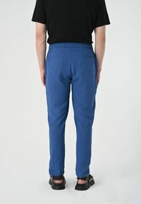 Blauwe broek met een elastische tailleband, voorzien van twee achterzakken en een slanke pasvorm. Gecombineerd met zwarte sandalen.