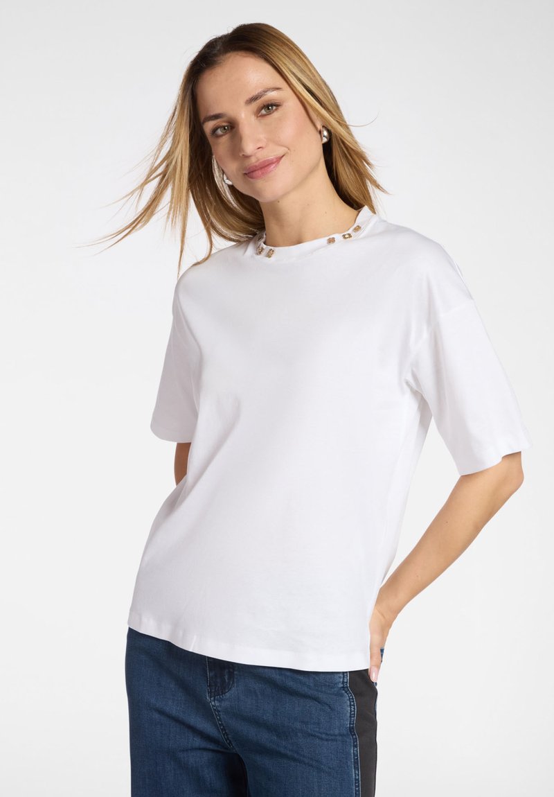 Vrouw met lang blond haar, gekleed in een wit shirt met korte mouwen en een decoratieve halslijn, en een blauwe spijkerbroek, glimlachend tegen een effen achtergrond.