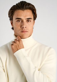 Krämfärgad polotröja med ribbad krage och muddar, mjuk textur, figurnära design, bärs av en person som håller handen mot hakan.