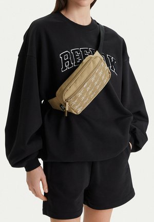 Osoba ubrana w czarną, oversize'ową bluzę z napisem "REFLEX" oraz czarne szorty, niosąca beżową torebkę na ramię z przednią kieszenią z siateczki.