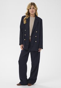 Blazer bleu marine adapté à double veste avec des boutons dorés, associé à un pantalon large assorti et un top en tricot beige. Chaussures marron texturées.
