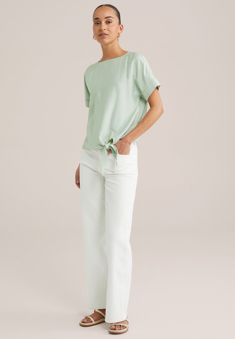 Heled roheline lühikeste varrukatega top, millel on vööl sõlme seotud, koos valgete laia säärega pükstega. Jalad kaunistatud pruuni sandaaliga.