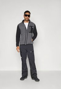 Pilkas ir juodas flisinis švarkas su užtraukiamu krūtinės kišeniu, derinamas su juodomis kelnėmis, kuriose yra šoninės užtrauktukai, ir tvirtais juodais batais.