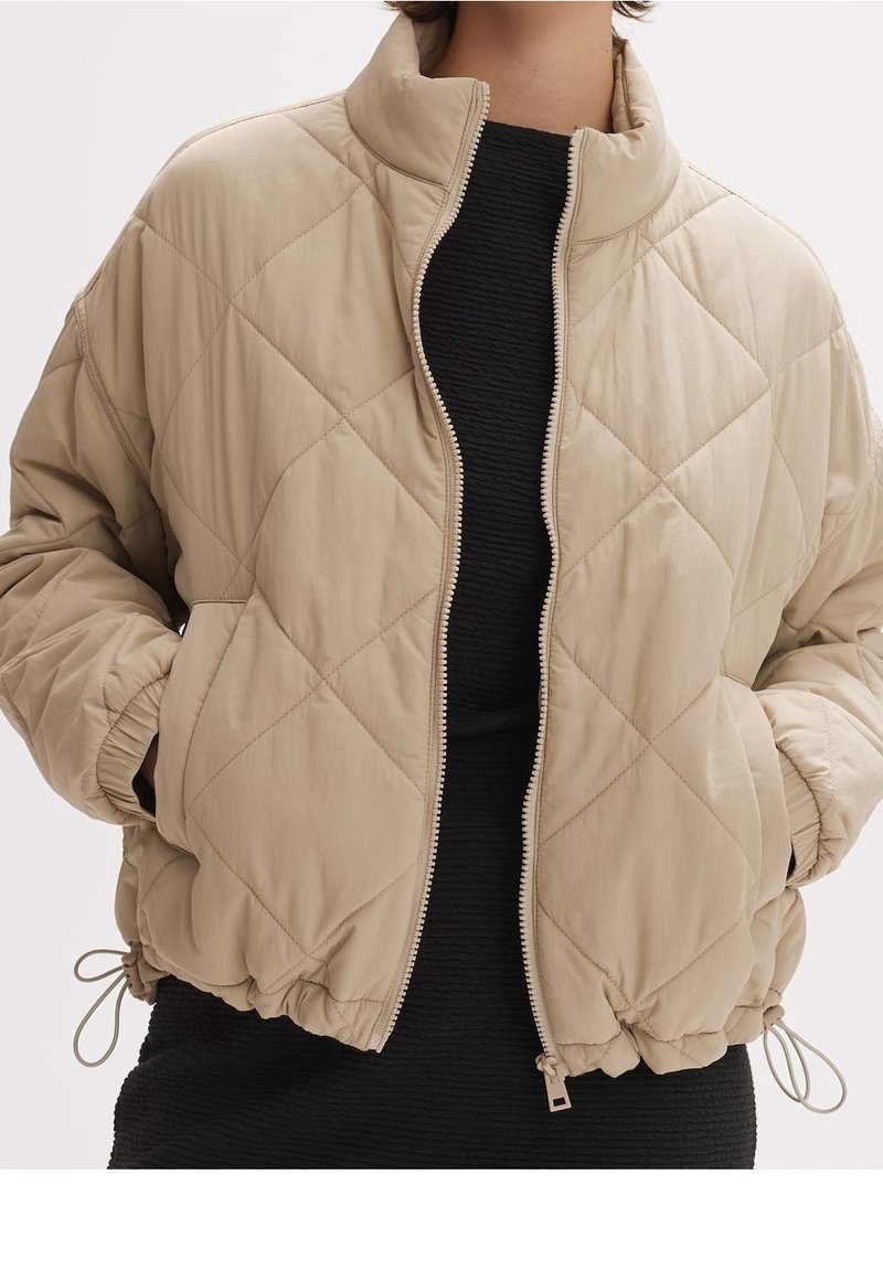 Beżowa pikowana kurtka z diamentowym przeszyciem, z przodem na zamek błyskawiczny, elastycznymi mankietami i ściągaczem u dołu noszona na teksturowanej czarnej sukience.