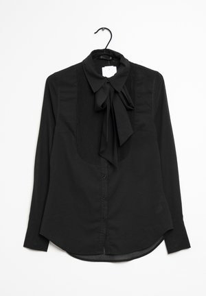 Blouse noire à manches longues avec fermeture boutonnée sur le devant, détail plissé sur la bavette et grand nœud noué au col, suspendue à un cintre noir.
