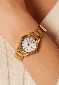 Horloge met een goudkleurige kast, een witte wijzerplaat, zwarte nummers en een datumaanduiding. Metalen armband met gepolijste schakels.