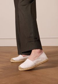 Weiße Leder-Espadrilles mit Jutesohlen, abgerundeten Zehen und beigen Akzenten. Kombiniert mit dunkel gefärbten Hosen mit Knopf am Saum. Holzfußboden im Hintergrund.