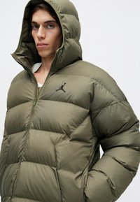 Young man wearing olive green padded jacket with hood, zipped halfway, showing a black Jumpman logo on the chest.