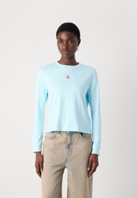 Hellblaues Langarmoberteil mit rundem Ausschnitt und kleinem roten "CK"-Logo auf der Brust, kombiniert mit hellen Denim-Jeans.