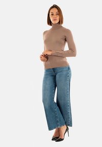 Beige coltrui top, nauwsluitend model, lange mouwen met knoopdetail. Lichtblauwe wijde pijpen jeans, denim textuur, gecombineerd met zwarte hoge hakken.