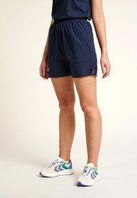 Marineblaue Shorts mit elastischem Bund, zwei Vordertaschen und einem pinken Logo-Detail. Kombiniert mit weißen und mehrfarbigen Sportschuhen.