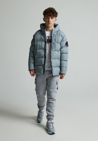 Doudoune gris clair avec capuche, dotée d'une fermeture éclair et de détails de logo. Associée à un pantalon de survêtement gris et des baskets grises.