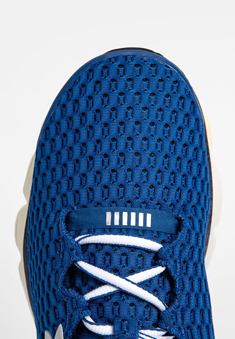 Zapatilla deportiva azul con una parte superior de malla texturizada, cordones blancos y una suela oscura sólida. Presenta acentos plateados minimalistas en la lengüeta.