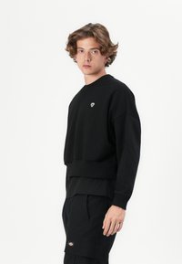 Black sweatshirt with a round neckline and ribbed hem. Features an embroidered logo on the chest. Soft texture, relaxed fit styling.