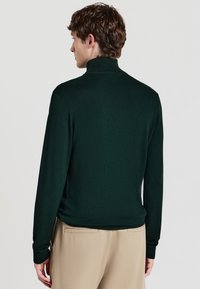 Dunkelgrüner Pullover mit hohem Kragen, schmaler Passform, langen Ärmeln und gerippten Bündchen. Das Material wirkt glatt, ohne Muster oder Verzierungen.