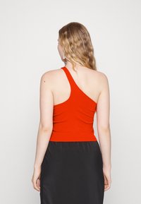 Top rosso a costine con design a spalla unica. Texture liscia, forma aderente e colore solido senza motivi o dettagli metallici.