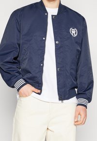 Navy bomber jacket made of smooth fabric, featuring a zip front, striped cuffs, and a logo design on the chest. Pockets on the sides.