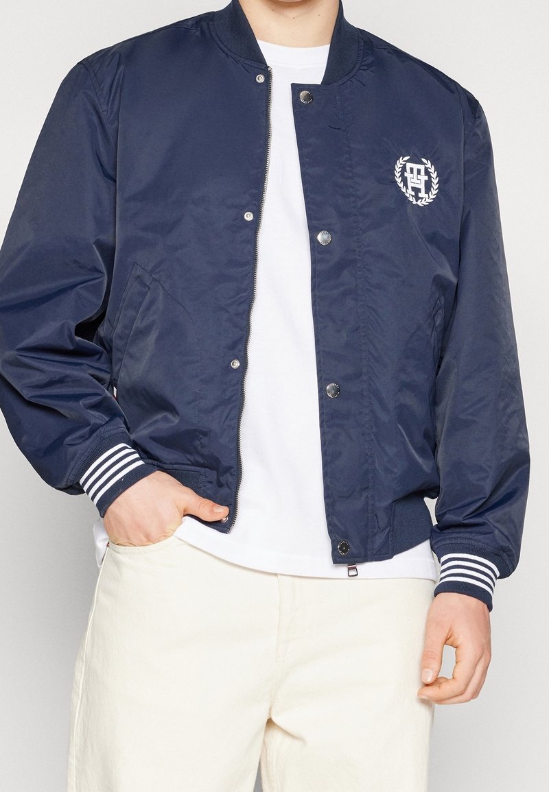 Navy bomber jacket made of smooth fabric, featuring a zip front, striped cuffs, and a logo design on the chest. Pockets on the sides.