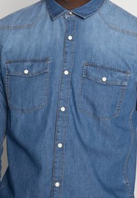 Camicia in denim con colletto button-down, di colore blu chiaro, due tasche sul petto e bottoni a pressione bianchi. Presenta dettagli di cucitura visibili.