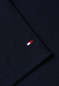 Gros plan sur un tissu bleu foncé avec un petit logo rectangulaire rouge, blanc et bleu marine sur le bord d'une manche pliée avec des coutures visibles.