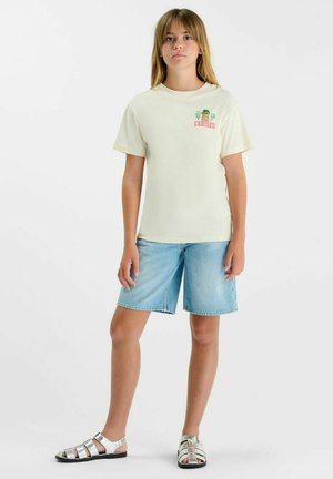 Jeune personne debout portant un t-shirt crème avec une petite impression colorée et un short en denim bleu clair avec des sandales argentées.