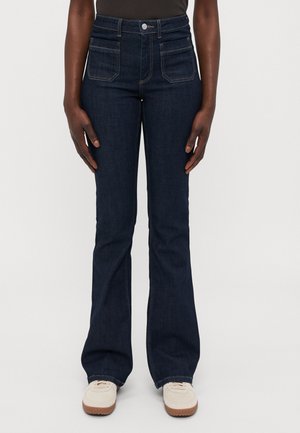 Jean flare en denim indigo foncé avec taille haute, dotés de poches avant, de surpiqûres contrastantes et d'une fermeture éclair. Associés à des chaussures claires.