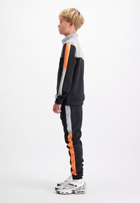 Zwart trainingspak met grijze en oranje strepen op de mouwen en broek. De outfit is gecombineerd met witte sneakers met zwarte accenten.