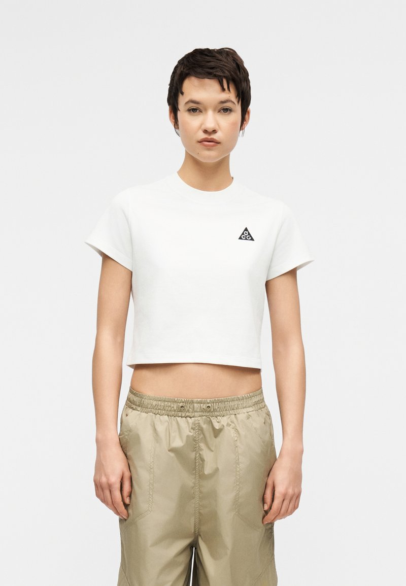 Young person with short dark hair wearing a white cropped T-shirt with a black triangle logo and beige drawstring pants, standing still.