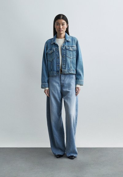 Γυναίκα στέκεται μπροστά σε απλό τοίχο, φοράει υπερμεγέθη μπλε τζιν μπουφάν, τζιν παντελόνι με ψηλή μέση και φαρδιά μπατζάκια, κρεμ ριμπ μπλούζα και μαύρα παπούτσια.