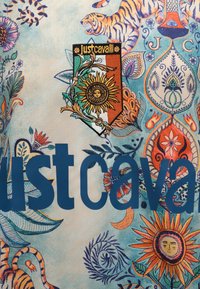 Υφασμα με λουλουδένιο και τίγρε μοτίβο, με το κείμενο «Just Cavalli» και ένα έμβλημα που απεικονίζει έναν ήλιο με πρόσωπο σε φόντο πράσινο, λευκό και πορτοκαλί.